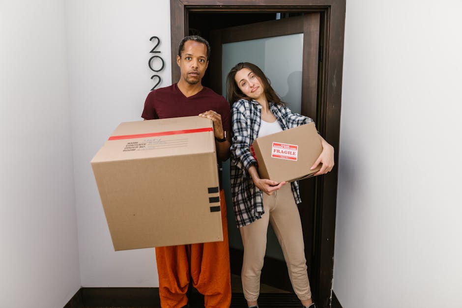 A man and a woman stand inside a doorway of a residential property, each holding cardboard boxes used for packing during a home relocation. The man, dressed in a maroon shirt and rust-coloured trousers, looks directly at the camera with a serious expression, while the woman, wearing a checkered shirt and beige pants, leans her head on the doorframe and gazes with a subtle smile. The boxes are sealed with packing tape; one has a red 'Fragile' sticker, indicating careful handling. The environment appears to be the entrance or hallway of a modern house, with the door open and showing a glimpse of the interior. The floor is dark and the walls are light, providing contrast. Visible are some partial house number details on the wall next to the doorframe. This setting reflects a typical loading process during a furniture transport or packing and moving service provided by Man with Van North Finchley, highlighting the preparation phase of a professional house removal within a local residential context, aligned with moving logistics and transportation services.
