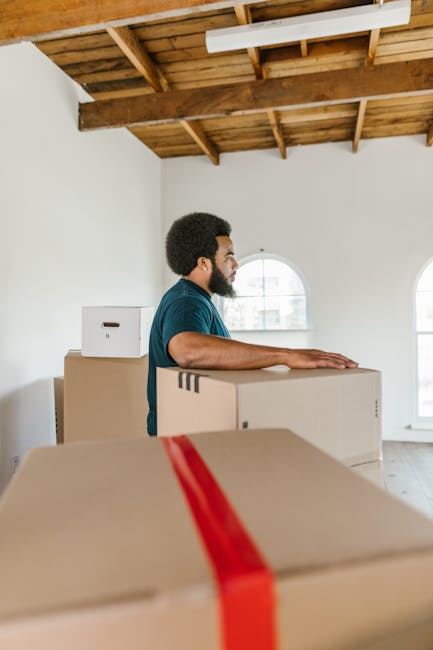 A man with dark skin, curly hair, and a beard, wearing a dark blue t-shirt, is seen inside a spacious, minimally furnished room with white walls and large, arched windows that let in natural light. He is standing behind a stack of large cardboard boxes, some of which are taped with red packing tape, indicating they are ready for or during a packing or home relocation process. The room features a wooden ceiling with exposed beams and a light fixture, and the floor appears to be a light-colored wood or laminate. The man has his hands resting on one of the boxes, possibly engaged in loading or organizing items as part of furniture transport or packing for a professional move. The setting suggests a house undergoing a move with visible materials typical of packing and moving processes, supported by the presence of multiple boxes that are characteristic of professional removals by Man with Van North Finchley, providing a context of home relocation or furniture transport.