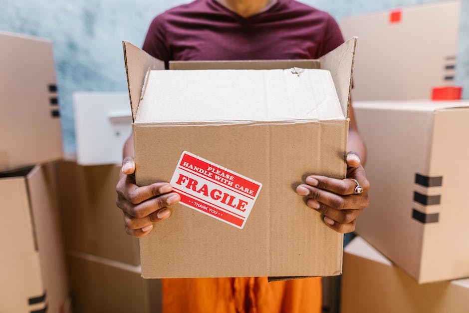 A man and a woman stand inside a doorway of a residential property, each holding cardboard boxes used for packing during a home relocation. The man, dressed in a maroon shirt and rust-coloured trousers, looks directly at the camera with a serious expression, while the woman, wearing a checkered shirt and beige pants, leans her head on the doorframe and gazes with a subtle smile. The boxes are sealed with packing tape; one has a red 'Fragile' sticker, indicating careful handling. The environment appears to be the entrance or hallway of a modern house, with the door open and showing a glimpse of the interior. The floor is dark and the walls are light, providing contrast. Visible are some partial house number details on the wall next to the doorframe. This setting reflects a typical loading process during a furniture transport or packing and moving service provided by Man with Van North Finchley, highlighting the preparation phase of a professional house removal within a local residential context, aligned with moving logistics and transportation services.
