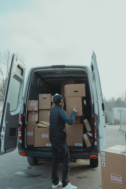 A man wearing a dark jacket, grey cap, and jeans is standing inside the open rear doors of a large white van, arranging and loading several cardboard boxes of various sizes, some with visible labels, into the vehicle. The boxes are made of brown corrugated cardboard, with some wrapped in clear plastic or secured with packing tape, and are stacked neatly on the van's floor, ready for home relocation or furniture transport. The scene is outdoors on a paved surface, with a cloudy sky overhead and a few trees or buildings visible in the distance. The open van doors reveal the empty interior, supported by loading equipment such as trolleys or straps, indicating an organized packing and moving process. This image is associated with professional removals services, such as those offered by Man with Van North Finchley, reflecting the logistics involved in house removals and moving procedures.