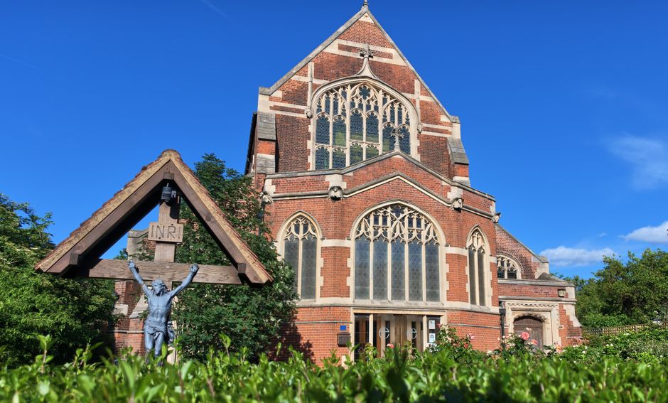 A large red-brick church with Gothic-style stained glass windows, including a prominent arched window on the front façade and additional smaller arched windows along the side. The church is surrounded by greenery, with trees and bushes visible around its perimeter. In the foreground, a small wooden shelter with a triangular roof displays a metal crucifix with a figure of Jesus Christ, mounted beneath a wooden cross marked with the inscription 'INRI.' The image appears to be taken on a bright, clear day with blue sky and sunlight illuminating the building and landscape. The scene suggests a peaceful, rural environment suitable for house removals or relocation services, with the church in view as part of local landmarks. The setting implies a calm neighborhood or village context, suitable for a professional home removal or furniture transport service like Man with Van North Finchley.