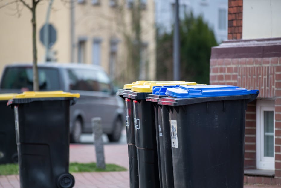A row of four large black wheelie bins with yellow, blue, and black lids positioned on a pavement outside a residential building, with a grey vehicle parked in the background. The bins are aligned in front of a brick wall adjacent to a white-framed basement window, and are ready for waste collection as part of a home relocation or household waste disposal process. Behind the bins, suburban residential buildings and trees are visible, indicating an urban or suburban environment. The scene is well-lit by daylight, emphasizing the materials and their placement, supporting the logistics involved in house removals and packing services offered by Man with Van North Finchley.