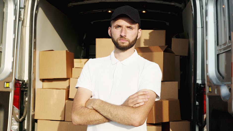 A man with a serious expression, wearing a black cap and a white short-sleeved shirt, stands inside the open rear area of a van filled with packed cardboard boxes of various sizes. The boxes are arranged on the floor and are secured for a home relocation or moving process. The van's interior is illuminated, with some natural light coming in from outside. The scene captures the loading or unloading phase of furniture transport, part of a professional removals service by Man with Van North Finchley, involving careful packing and moving logistics in preparation for or during a house relocation.