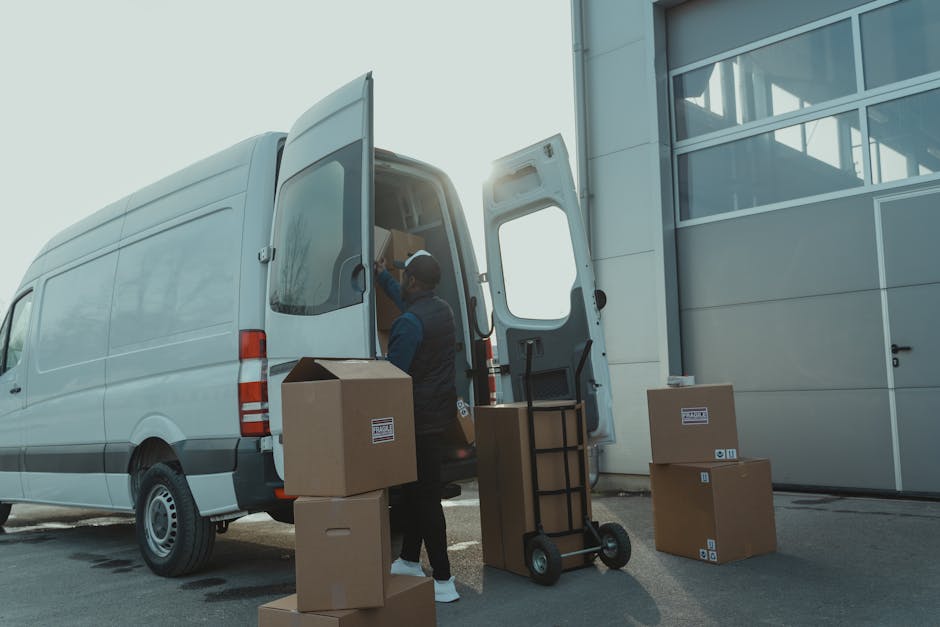 A professional mover from Man with Van North Finchley is loading a white panel van with cardboard boxes and packing supplies outside a modern building with large glass windows. The mover, dressed in dark clothing, is seen placing boxes into the open rear doors of the van, which is parked on pavement, with some boxes resting on the ground nearby. A small hand truck or dolly with additional packing materials is positioned close to the vehicle, ready for the home relocation process. The scene captures the loading process during daylight, with the sun slightly visible behind the building, emphasizing the logistical aspect of furniture transport and packing in preparation for a national or local move, reflecting efficient removal services offered by [COMPANY_NAME].