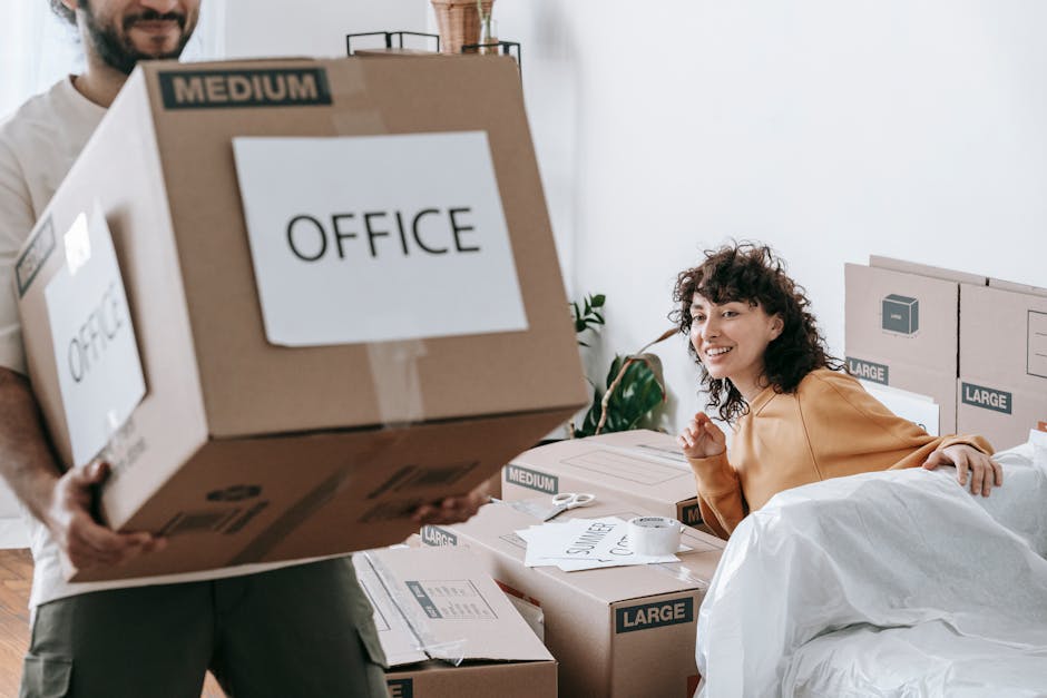 A man with dark, curly hair and a beard, dressed in a beige shirt, is lifting a large cardboard box labeled 'OFFICE' with a 'MEDIUM' weight indicator, during a home or office relocation process inside