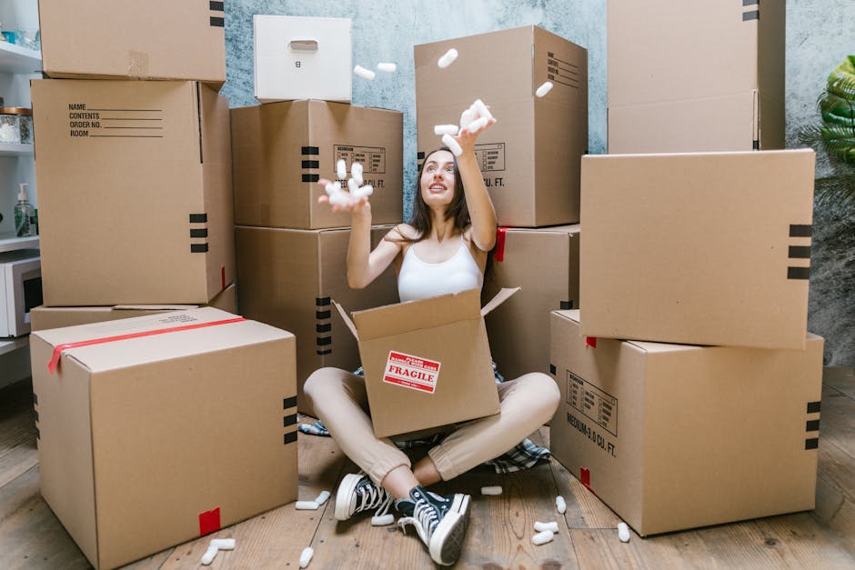 A young woman with long, curly hair and a neutral expression is standing indoors near a doorway, holding two cardboard boxes, one stacked on top of the other. The top box has the word 'toys' handwritt